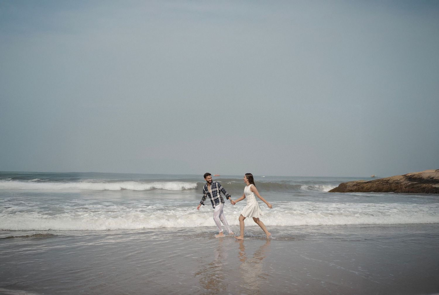 A couple joyfully runs along a sandy beach with ocean waves crashing behind them on a cloudy day.