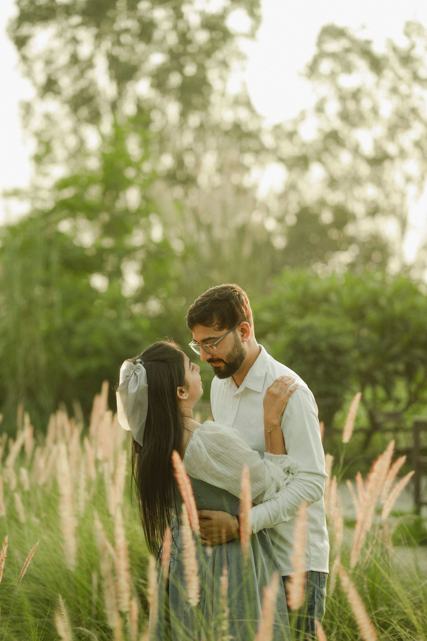 Couple embracing romantically in a lush green park with tall grass and trees in warm golden light.