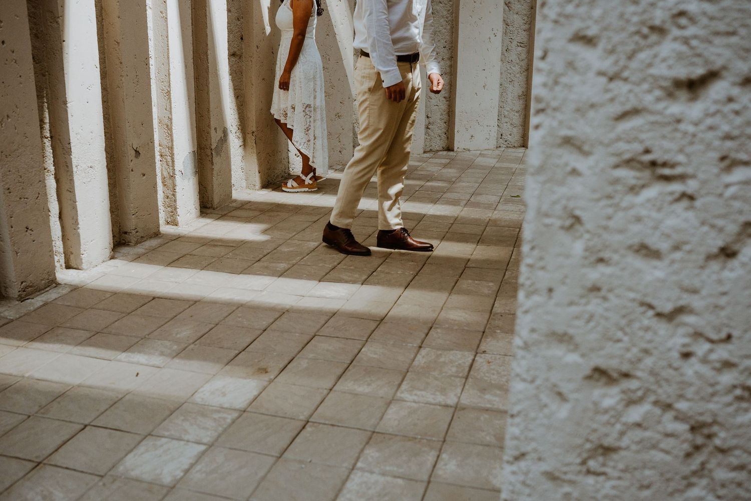 Couple walking through a sunlit colonnade with beige tiles and white stone pillars.