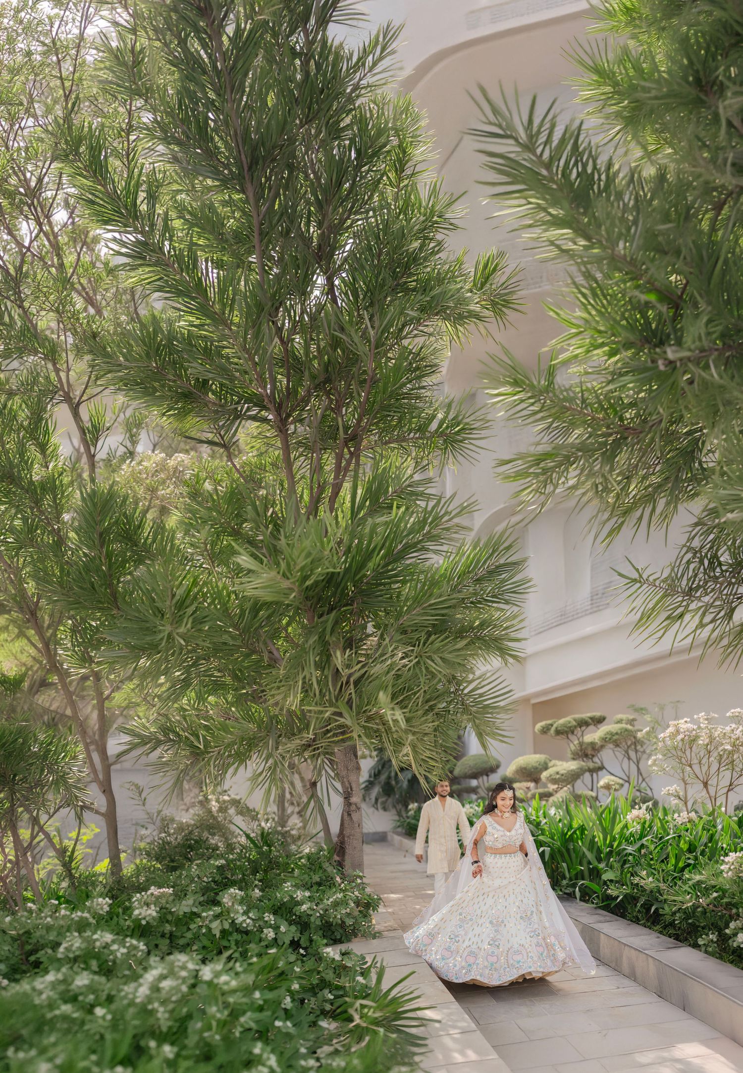A couple in wedding attire walks hand-in-hand through a lush garden pathway beside an elegant white building.