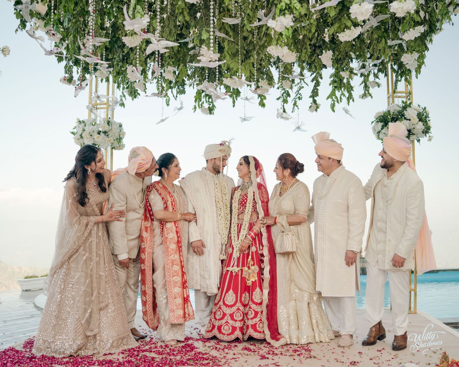A wedding party in traditional South Asian attire poses on a beach with ocean views and floral decorations.