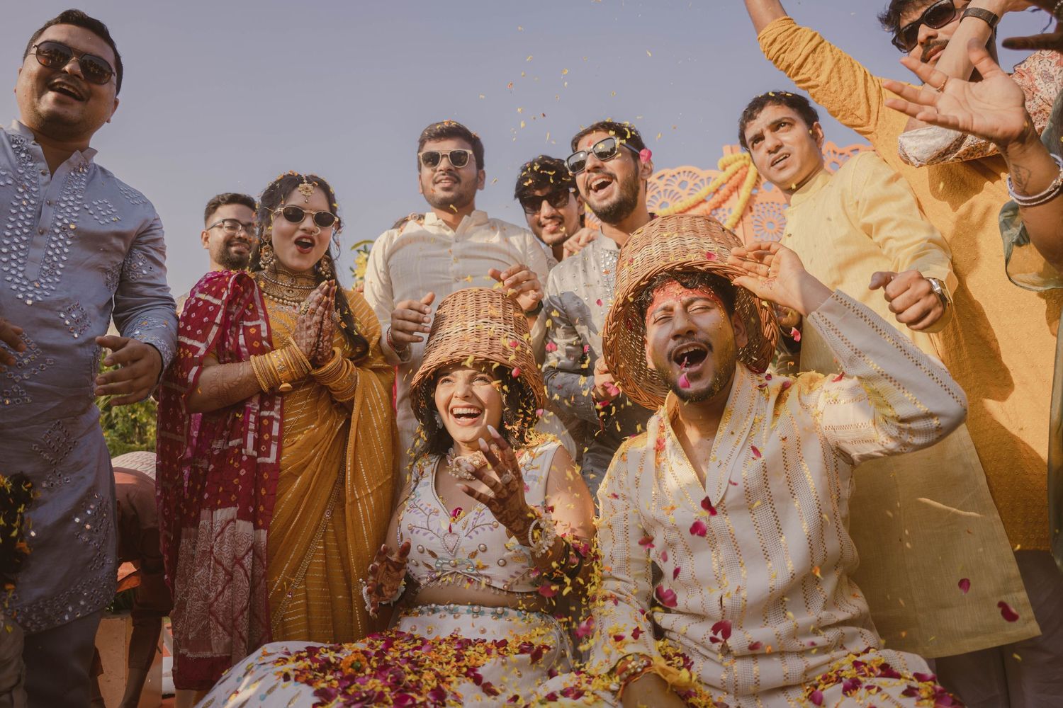 Joyful wedding celebration with guests throwing colorful flower petals in golden sunlight at outdoor ceremony.