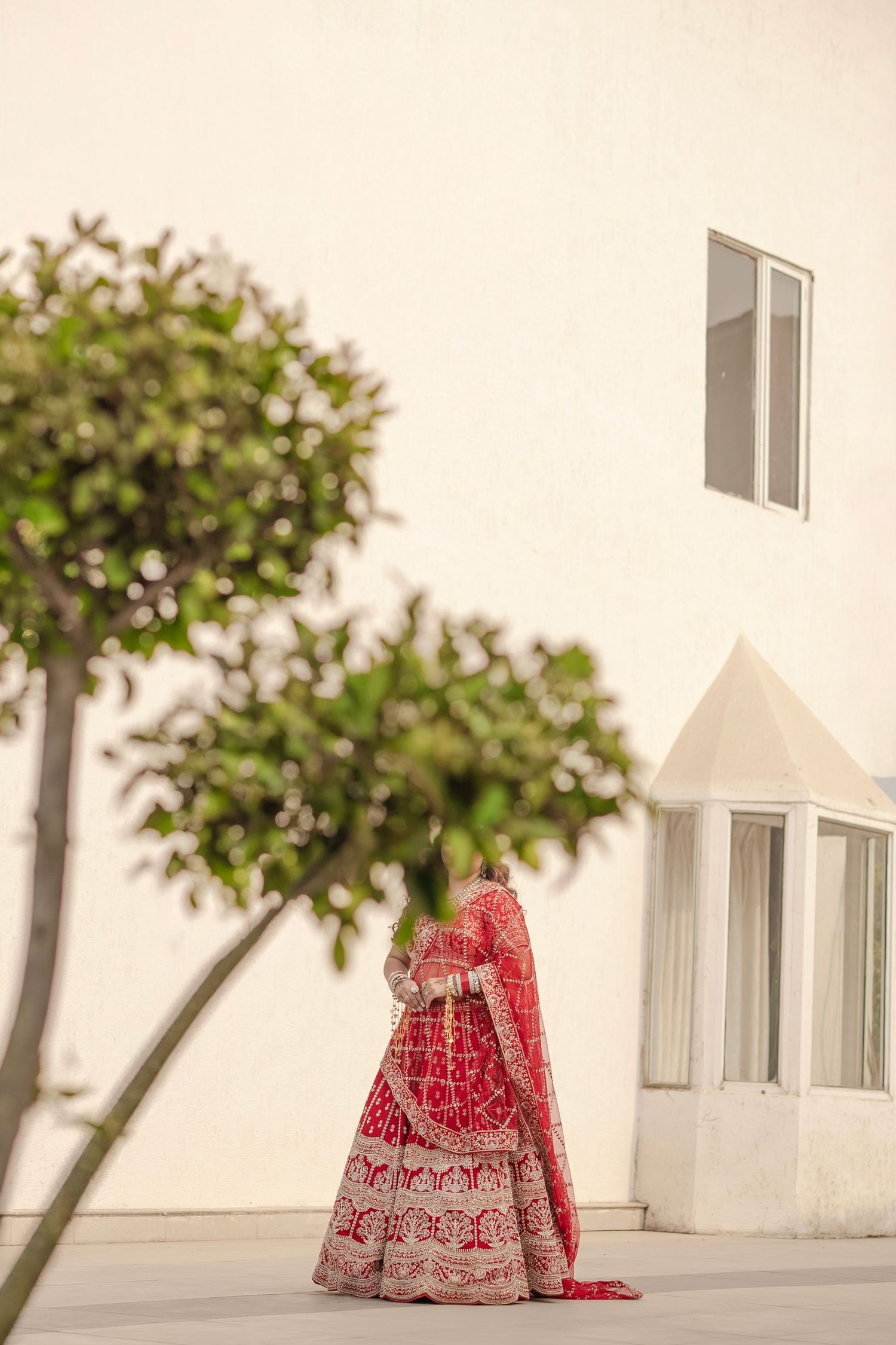Bride in red embroidered lehenga with dupatta poses outdoors near white building and green trees.