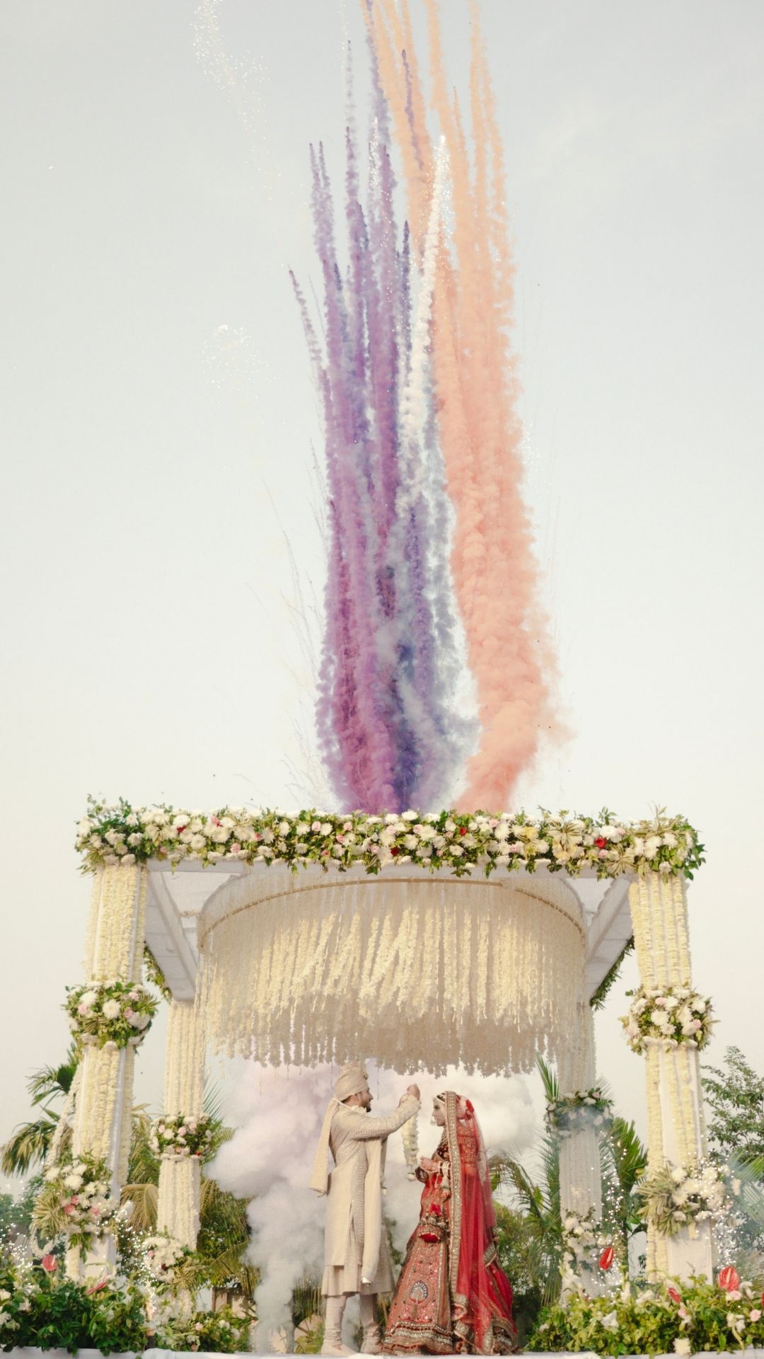 Indian wedding couple under floral mandap with colorful smoke trails from aerobatic display in sky.