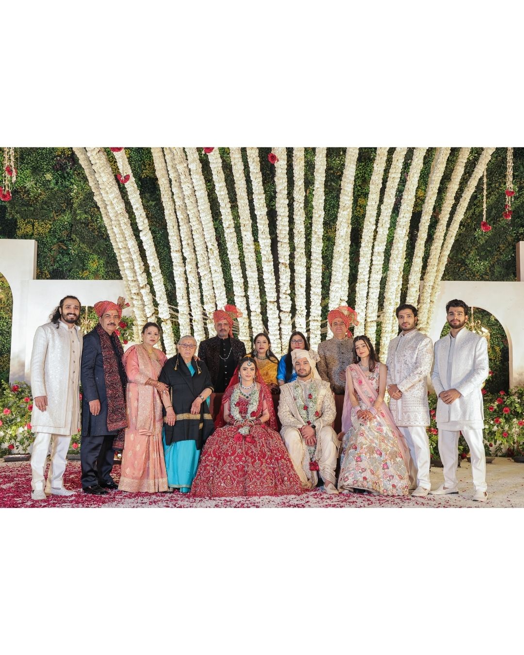 Indian wedding group photo with bride in red lehenga and groom in sherwani, surrounded by family under floral arch.