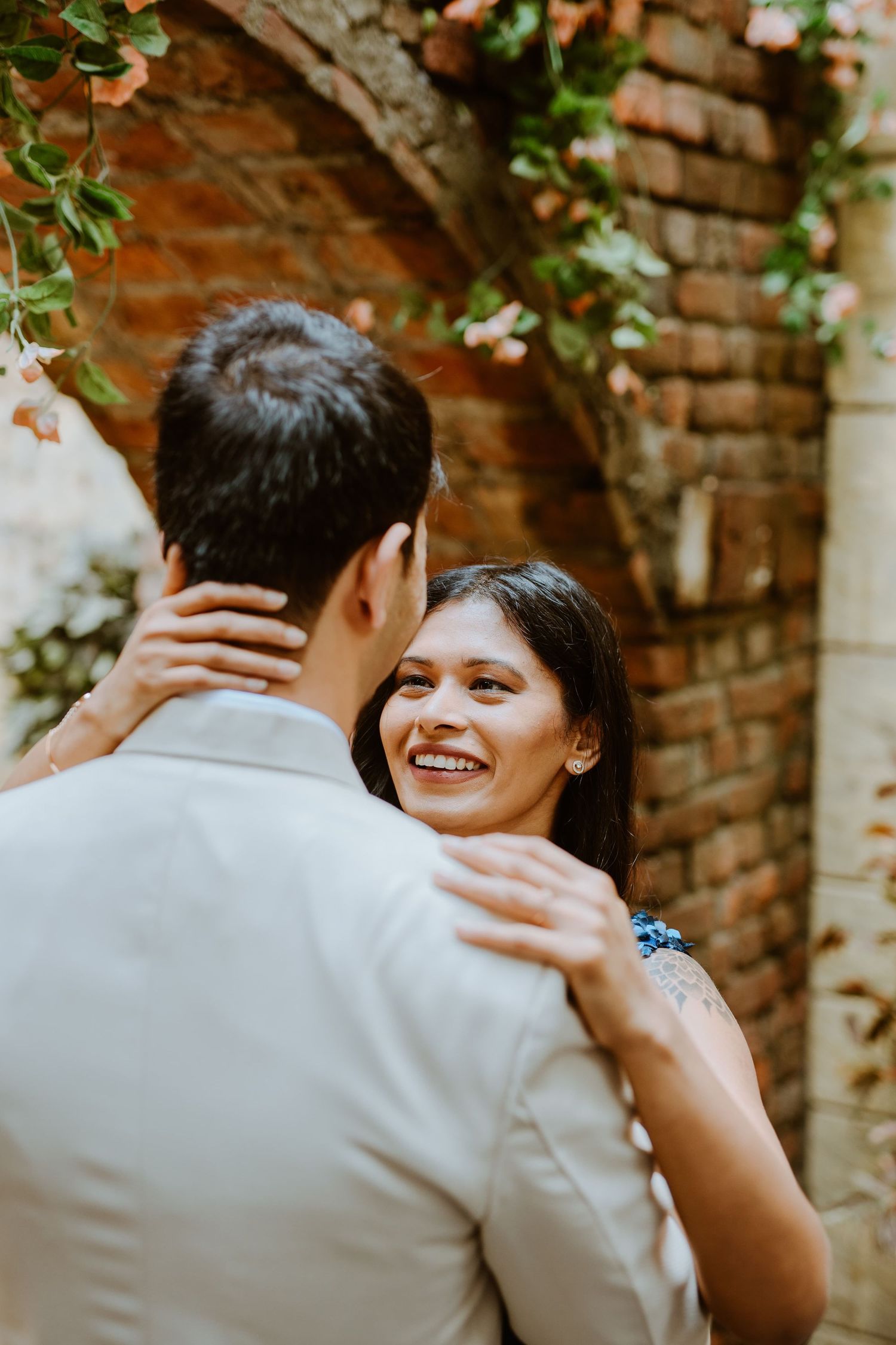 Smiling woman embraces man outdoors near rustic brick arch with flowers, sharing a romantic moment.