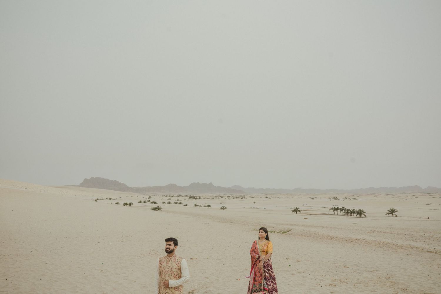 A couple in traditional South Asian attire standing apart in a vast sandy desert landscape under a hazy sky.