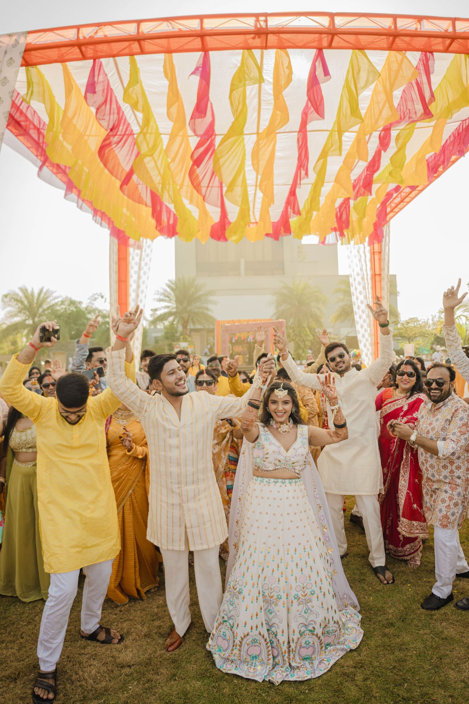 Indian wedding couple celebrates under colorful yellow and pink fabric canopy surrounded by joyful guests in traditional attire.