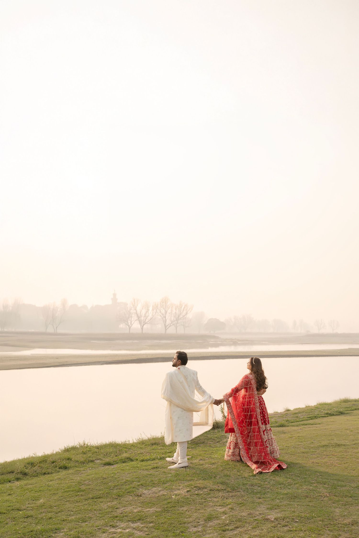 Indian couple in traditional attire holding hands by a misty riverside, groom in white sherwani, bride in red lehenga.