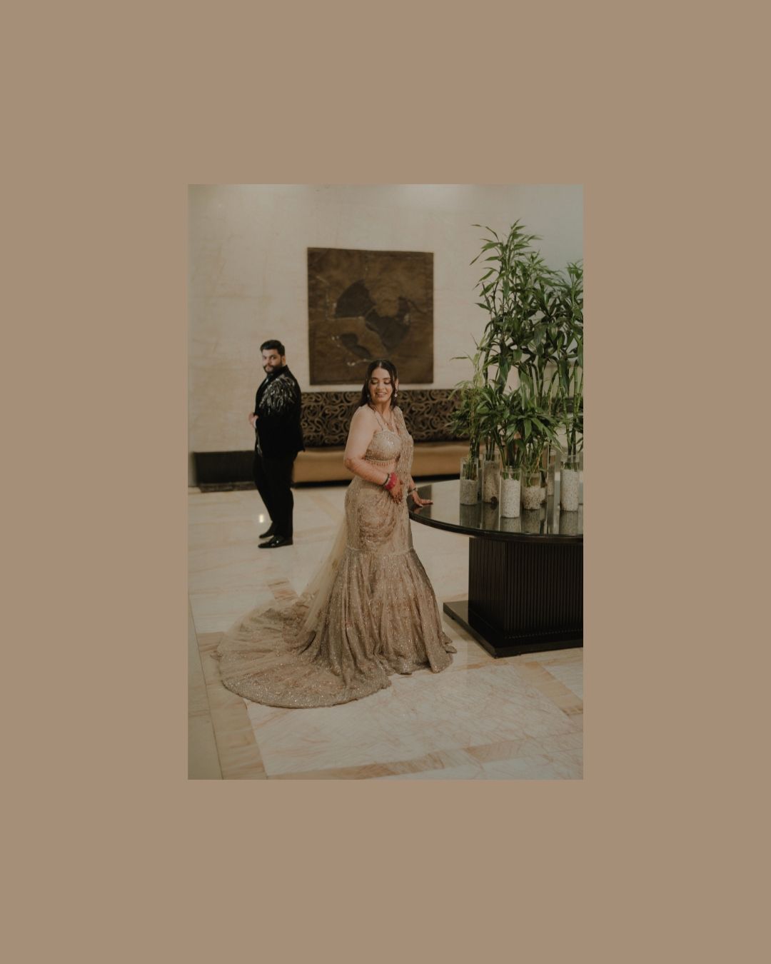 Woman in elegant gold lehenga poses in luxury hotel lobby while man in black stands in background.