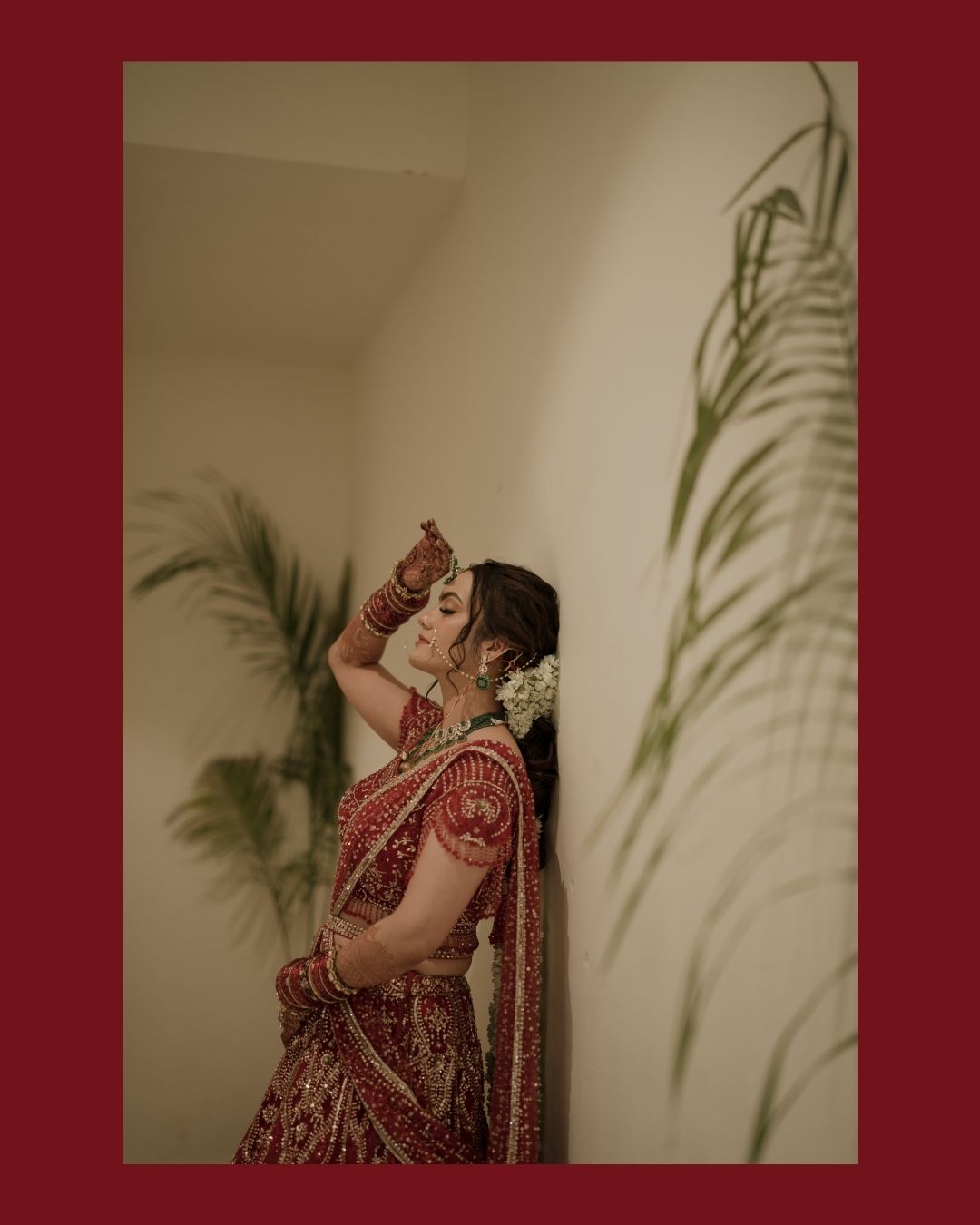 A bride in a red embroidered lehenga with gold jewelry and white flowers in her hair poses elegantly indoors.