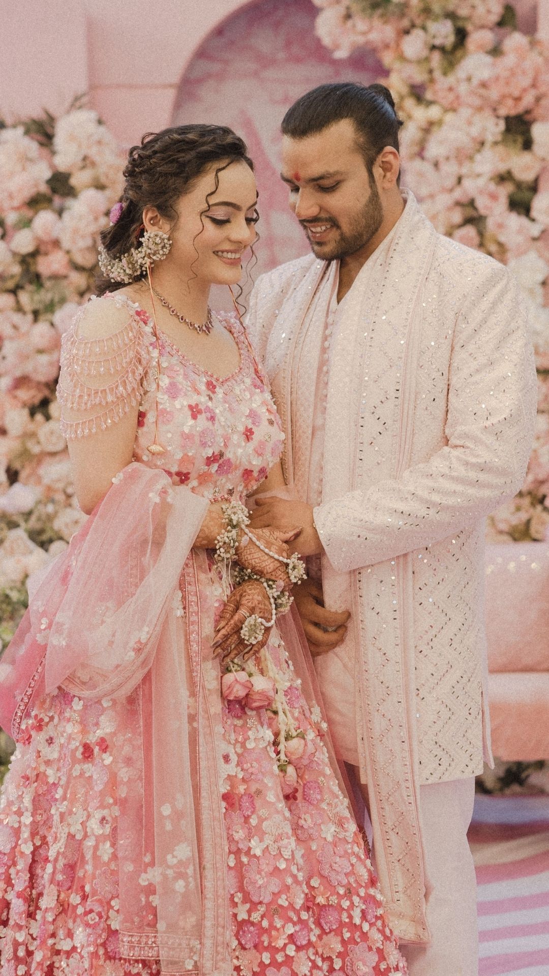 A couple in traditional Indian wedding attire, bride in pink floral lehenga and groom in white sherwani, posing before floral backdrop.