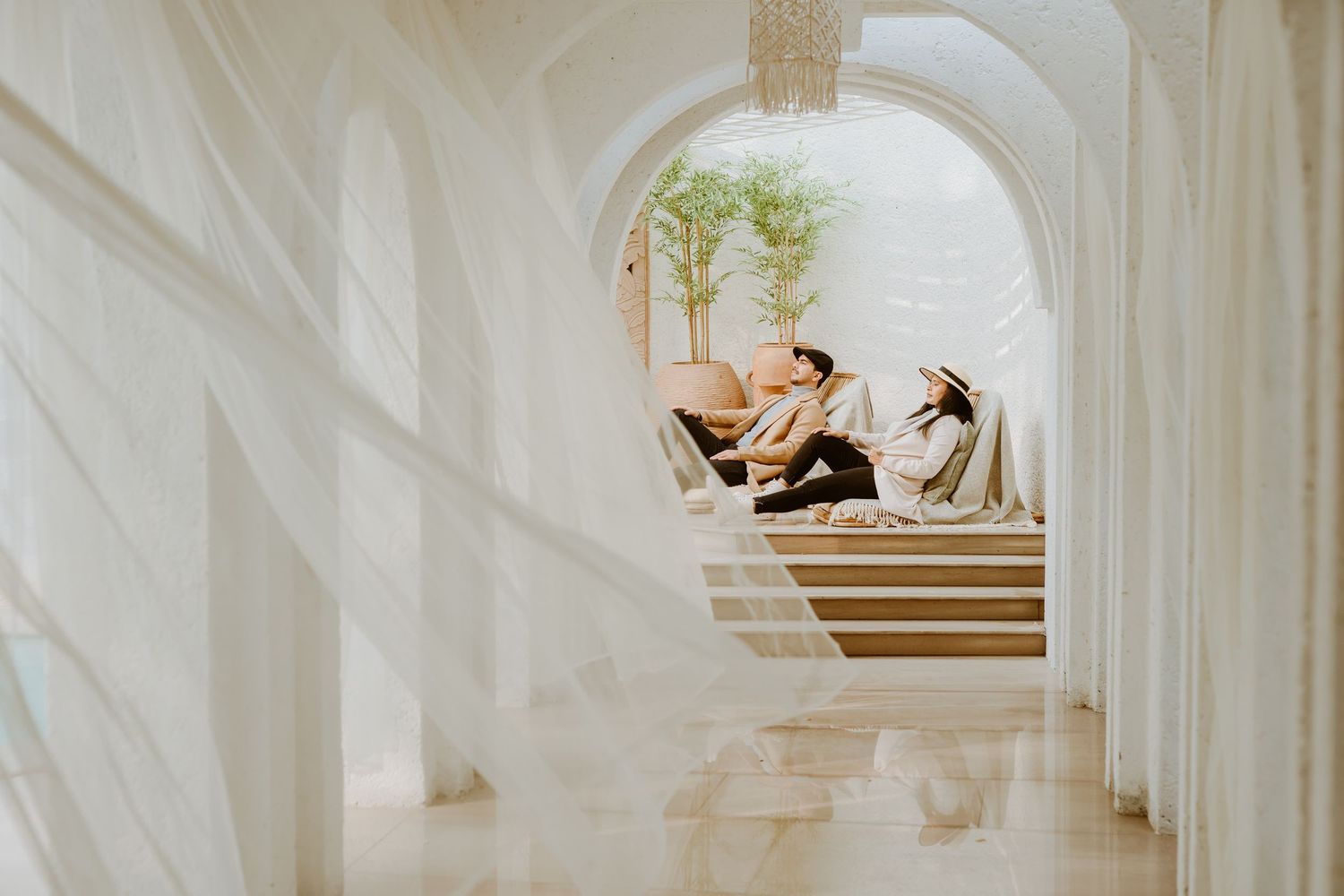Two people relaxing on lounge chairs in a serene white arched spa corridor with soft natural lighting.