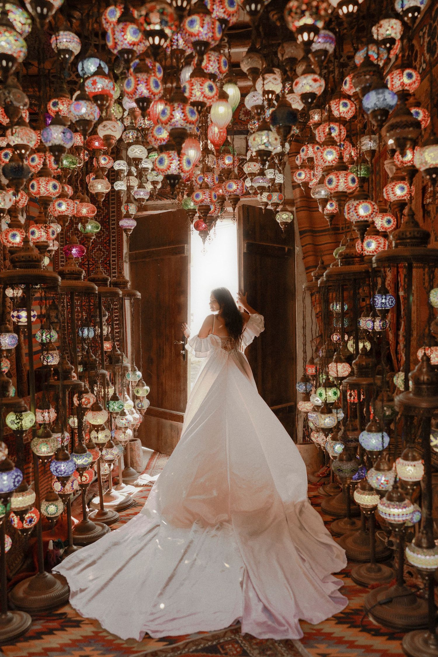 Bride in white gown stands among colorful Turkish mosaic lanterns in an ornate doorway glowing with warm light.