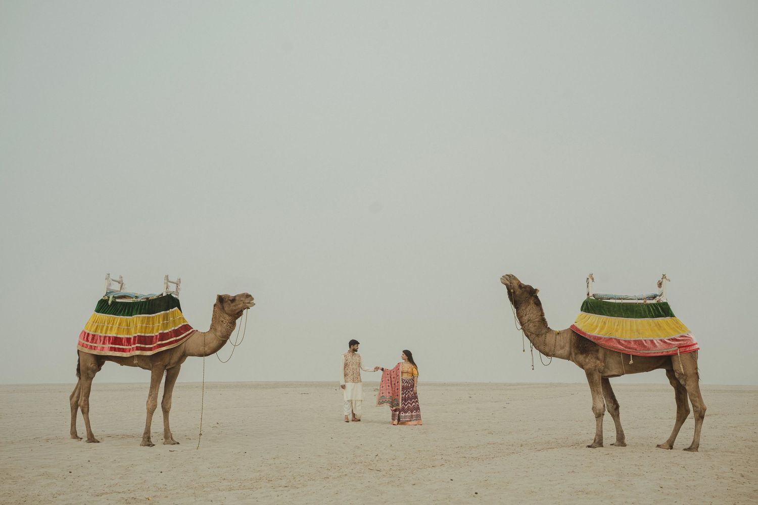 Couple standing between two decorated camels in a vast desert landscape under a hazy sky.