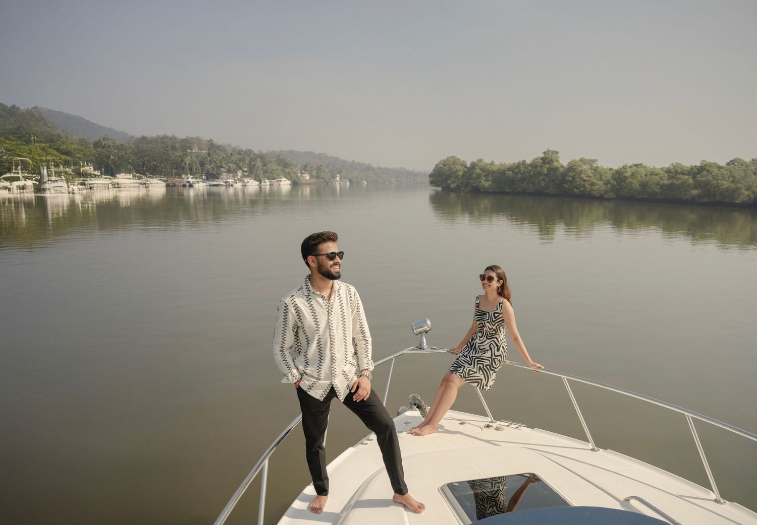 Stylish couple posing on a luxury yacht bow on a calm river surrounded by lush green trees.
