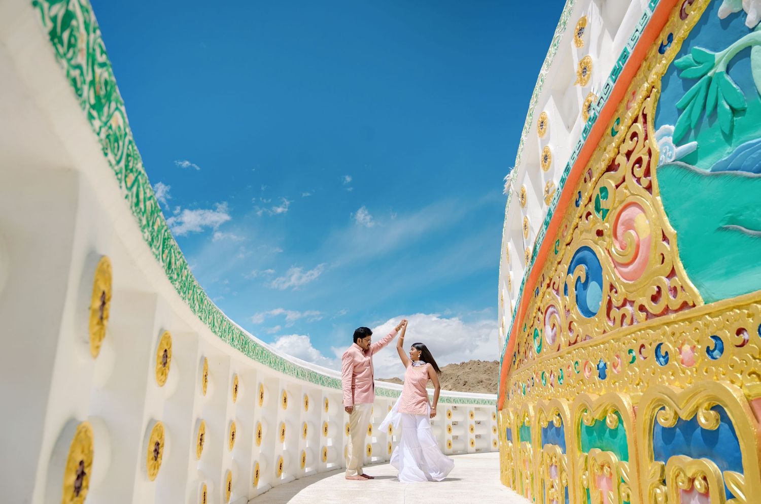 Couple dancing at ornate white Buddhist temple with colorful murals under vivid blue sky.