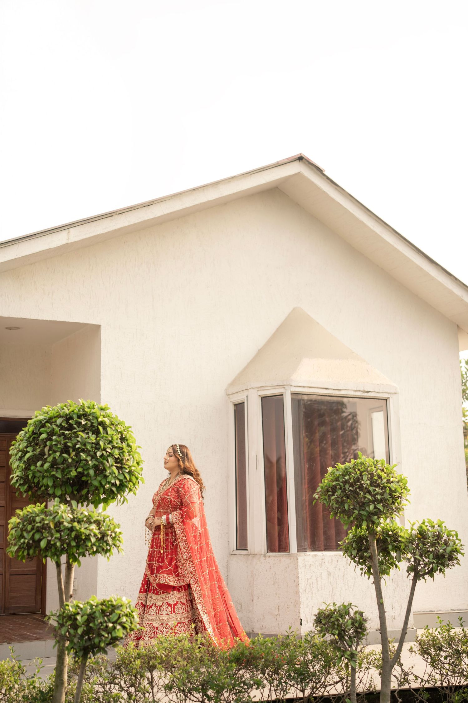 Bride in red lehenga standing outside white house with trimmed green trees.