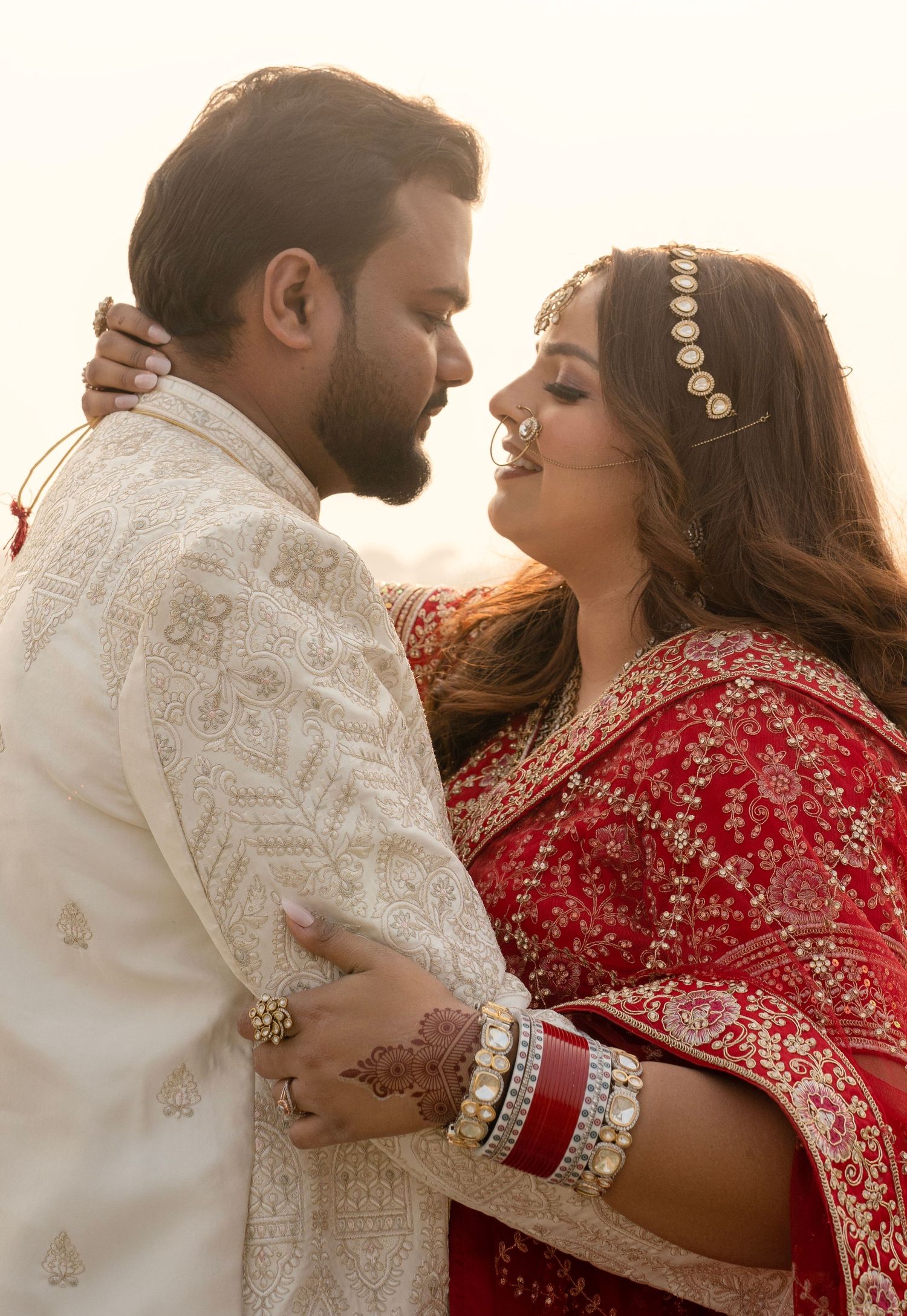 Couple in traditional Indian wedding attire sharing romantic moment, bride in red lehenga with gold jewelry, groom in white sherwani.