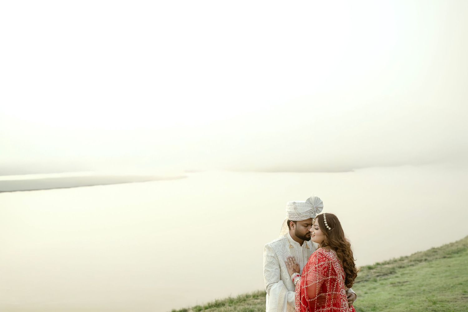 Couple in traditional Indian wedding attire sharing a romantic moment by a serene riverside.