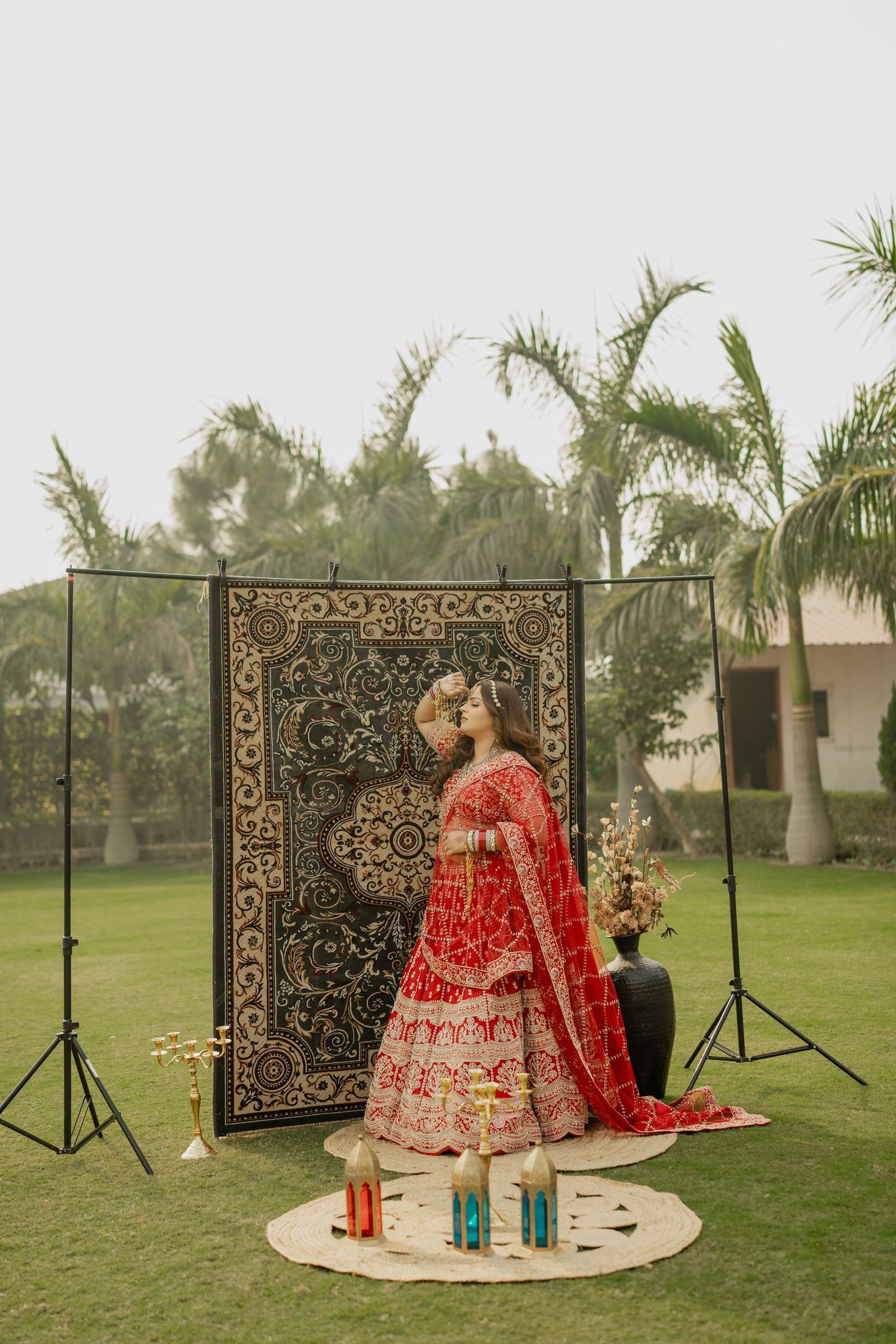 Bride in red bridal lehenga poses outdoors before ornate black tapestry backdrop with palm trees and colorful lanterns.