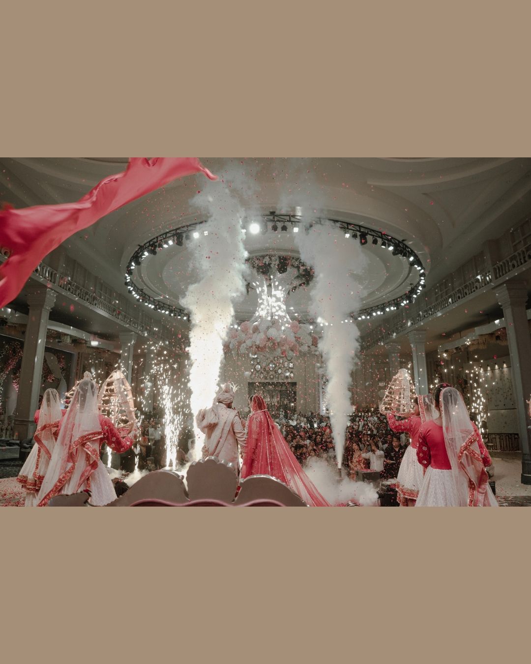 Grand Indian wedding ceremony with fireworks, smoke effects, and guests in red and white attire under an ornate chandelier.