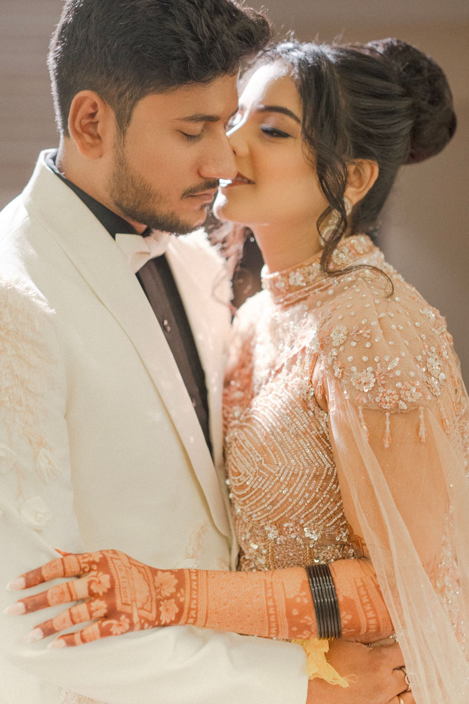 A South Asian bride and groom share an intimate moment, bride wearing embroidered lehenga with mehndi, groom in white tuxedo.