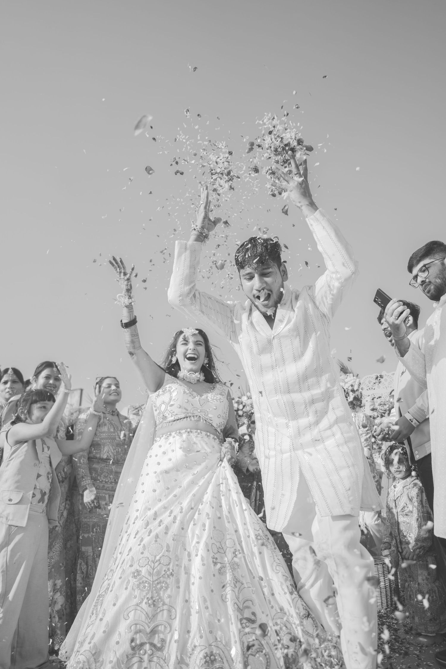 Black and white photo of a joyful couple in traditional Indian wedding attire celebrating with confetti and guests.