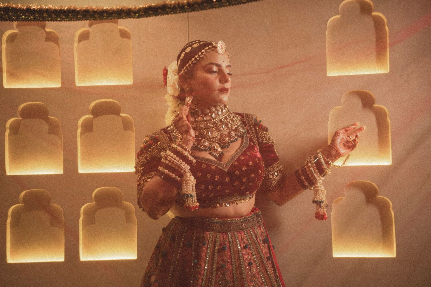 Woman in ornate red bridal lehenga with gold jewelry poses gracefully against decorative arch wall backdrop.