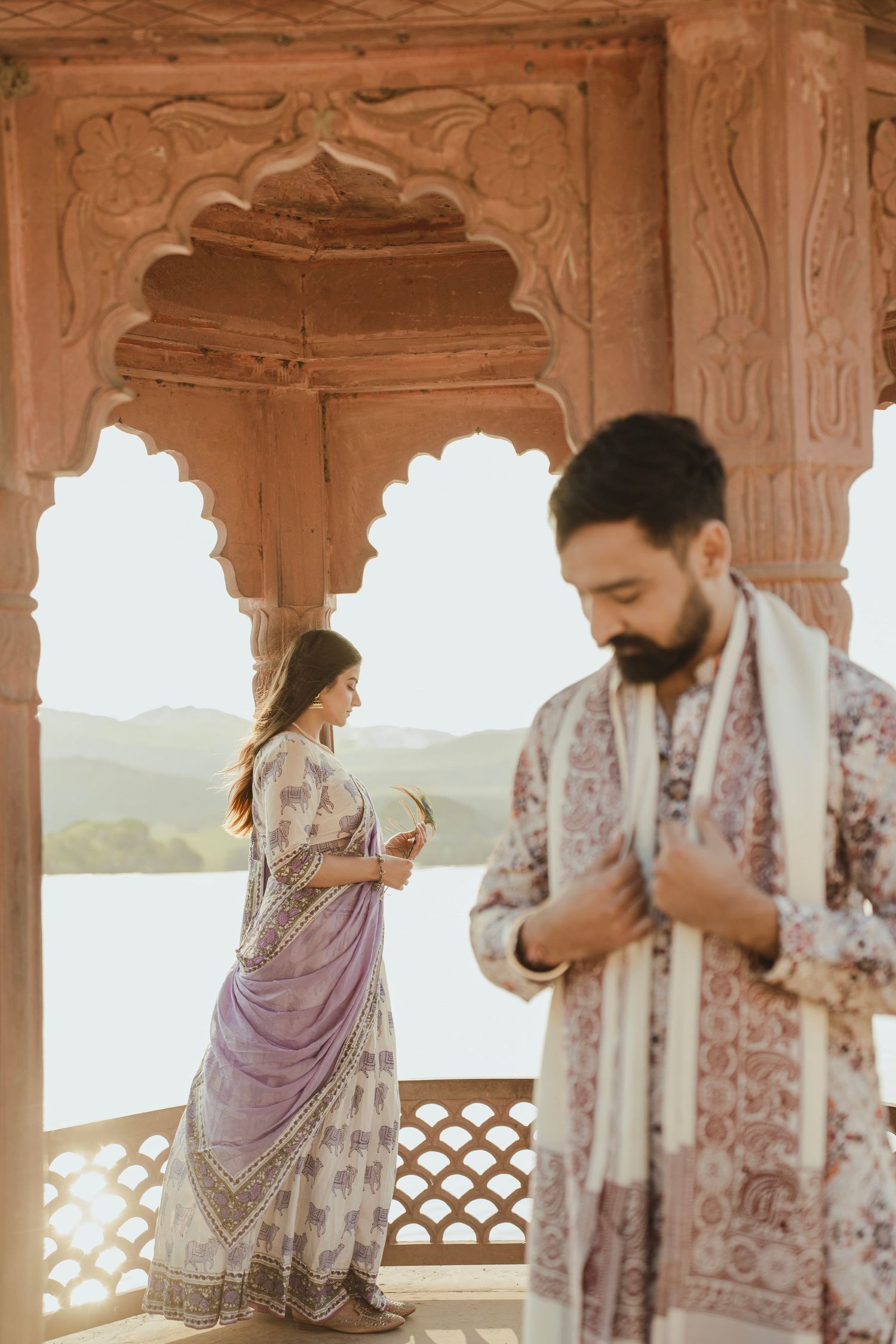 Couple in traditional Indian attire at ornate red sandstone arched pavilion overlooking scenic lake and hills.
