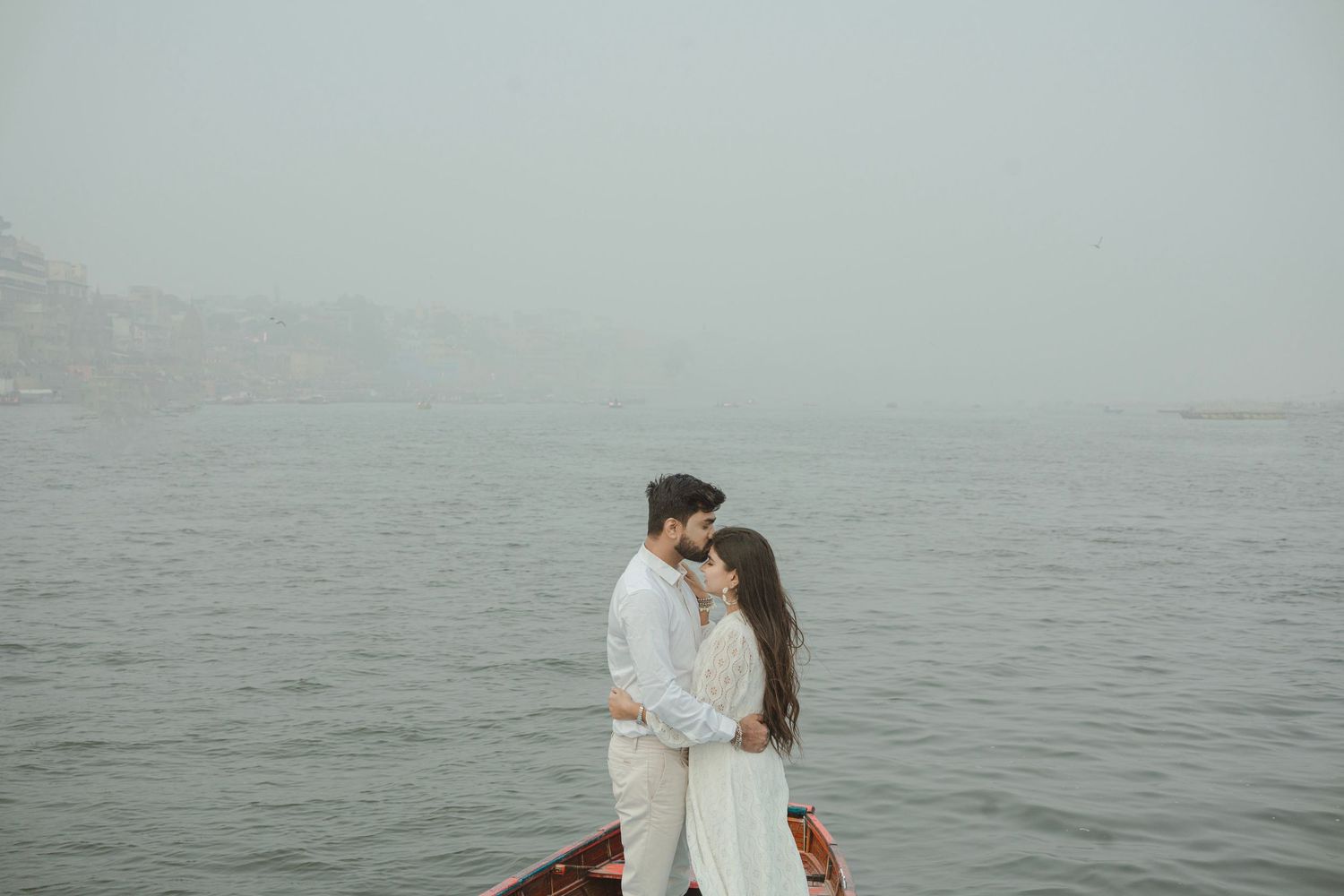 Romantic couple embracing on a boat in misty foggy water, wearing white outfits, with hazy cliffs in background.