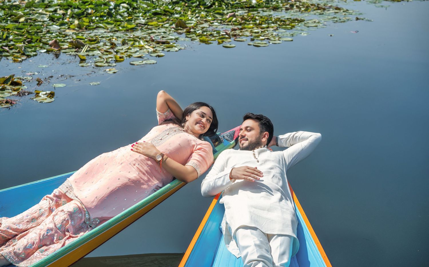 Couple in traditional Indian attire relaxing on a colorful boat surrounded by lily pads on a serene lake.