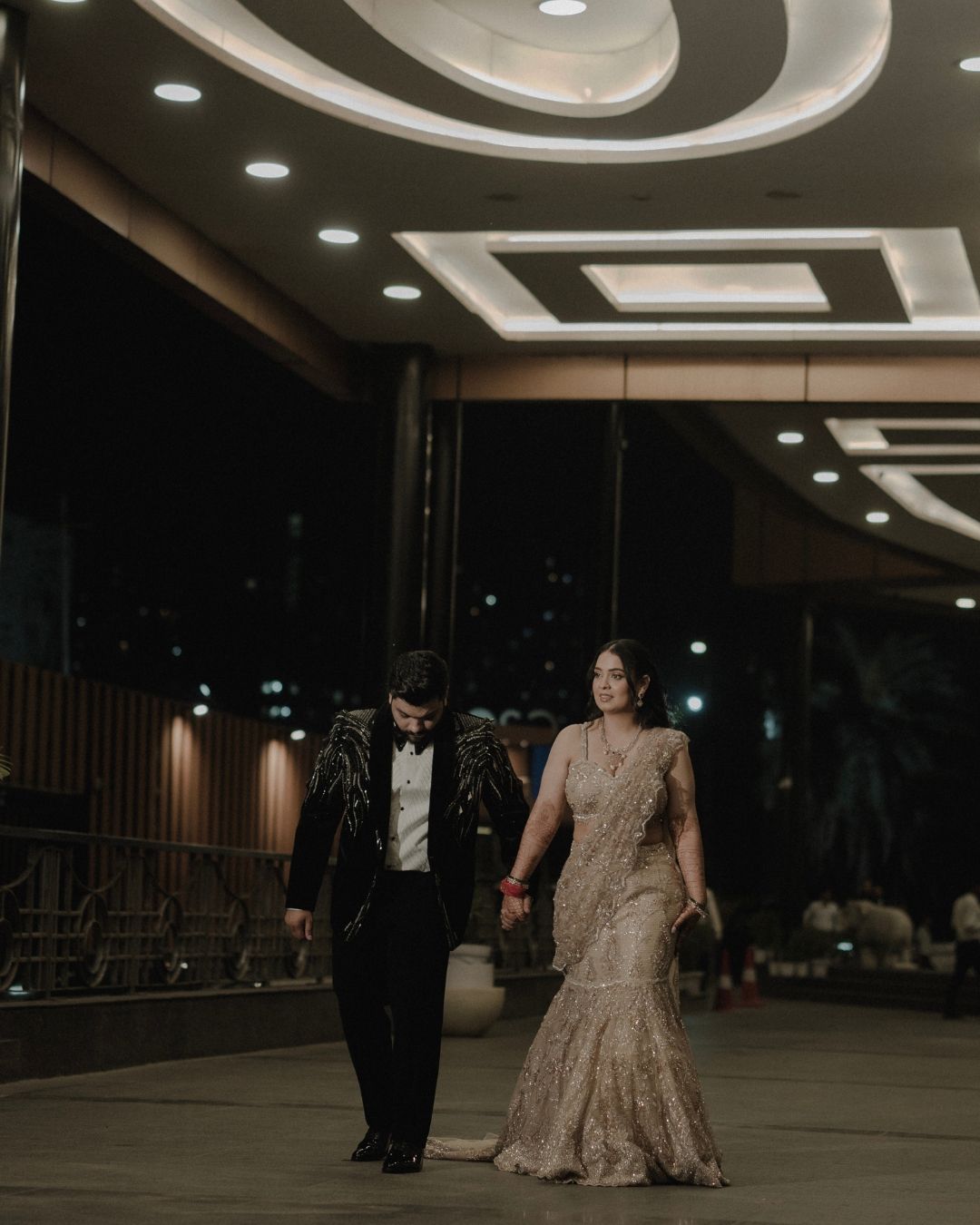 Elegant couple walking hand in hand at night, woman in gold saree and man in black tuxedo, under ornate ceiling lights.