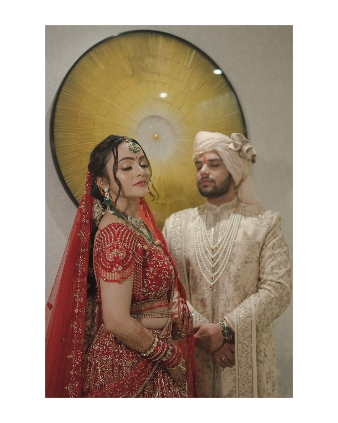Indian bride in red lehenga and groom in ivory sherwani pose together at a traditional Hindu wedding ceremony.