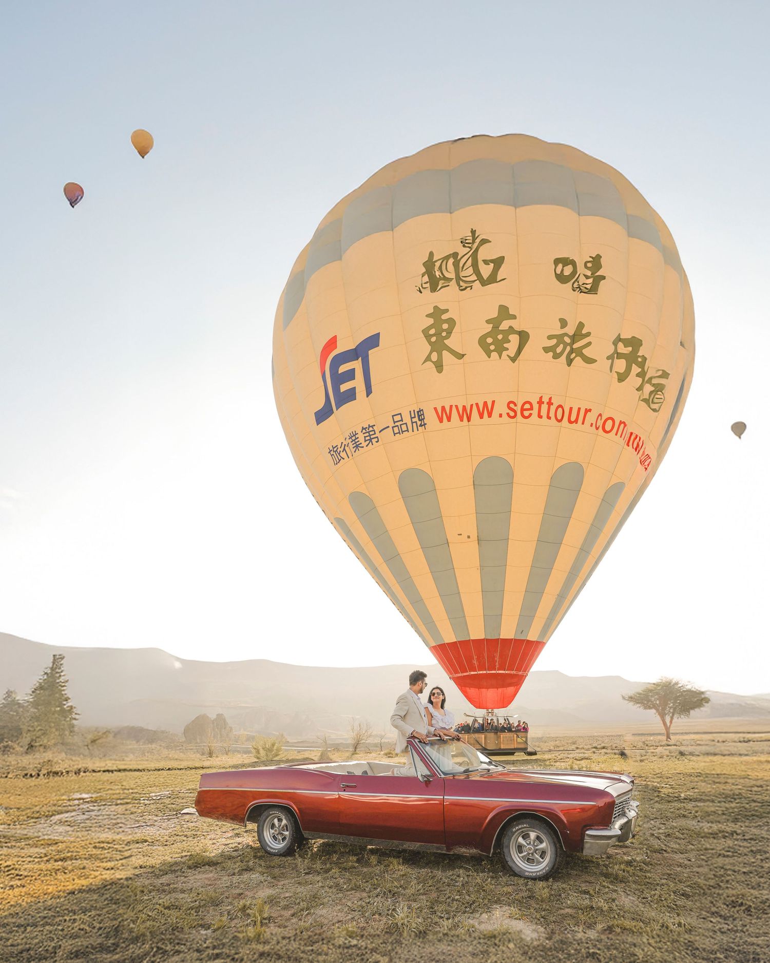 Couple posing beside a red convertible car with a large SET Tour hot air balloon in a scenic open field.