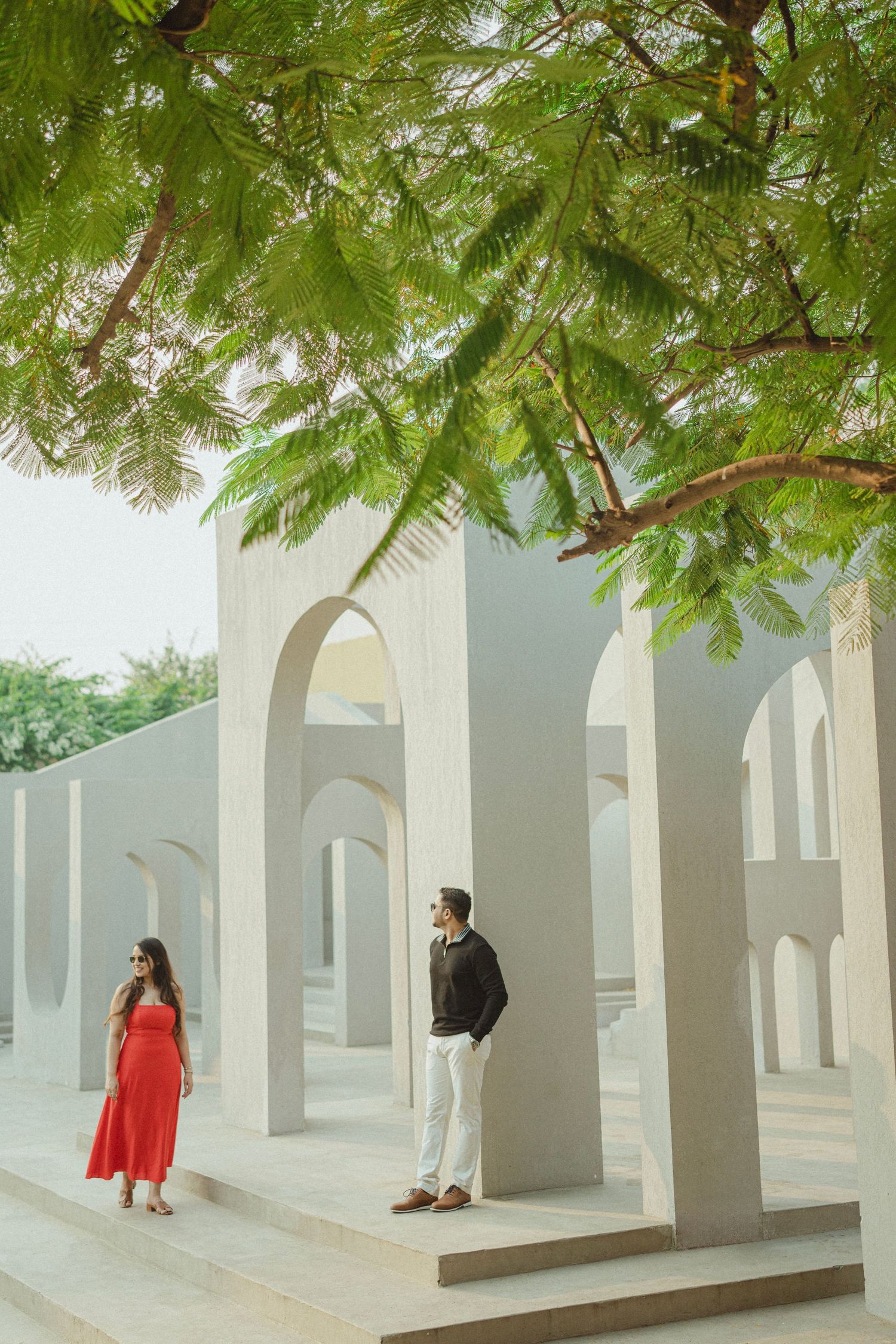 Couple posing near white arched columns under a green tree canopy, woman in red dress, man in black jacket.