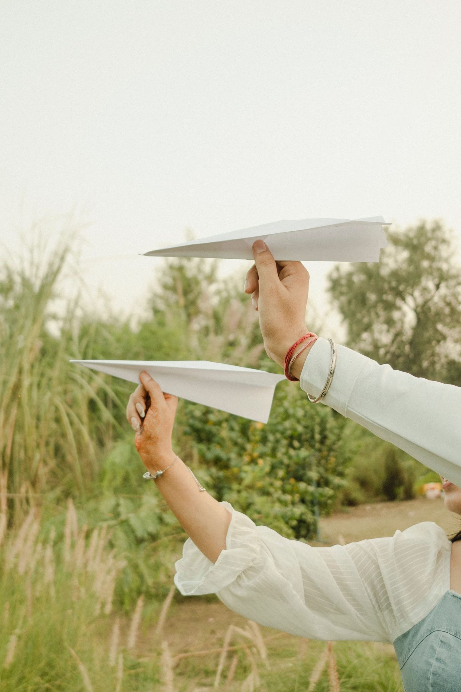 Two hands holding white paper airplanes outdoors with green trees and tall grass in the background.