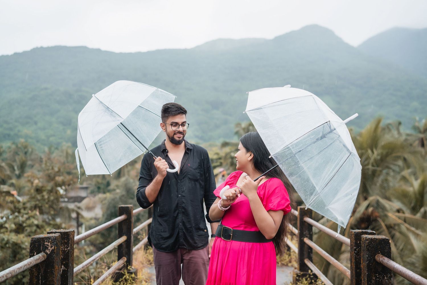 Couple with clear umbrellas posing together on a scenic mountain overlook.