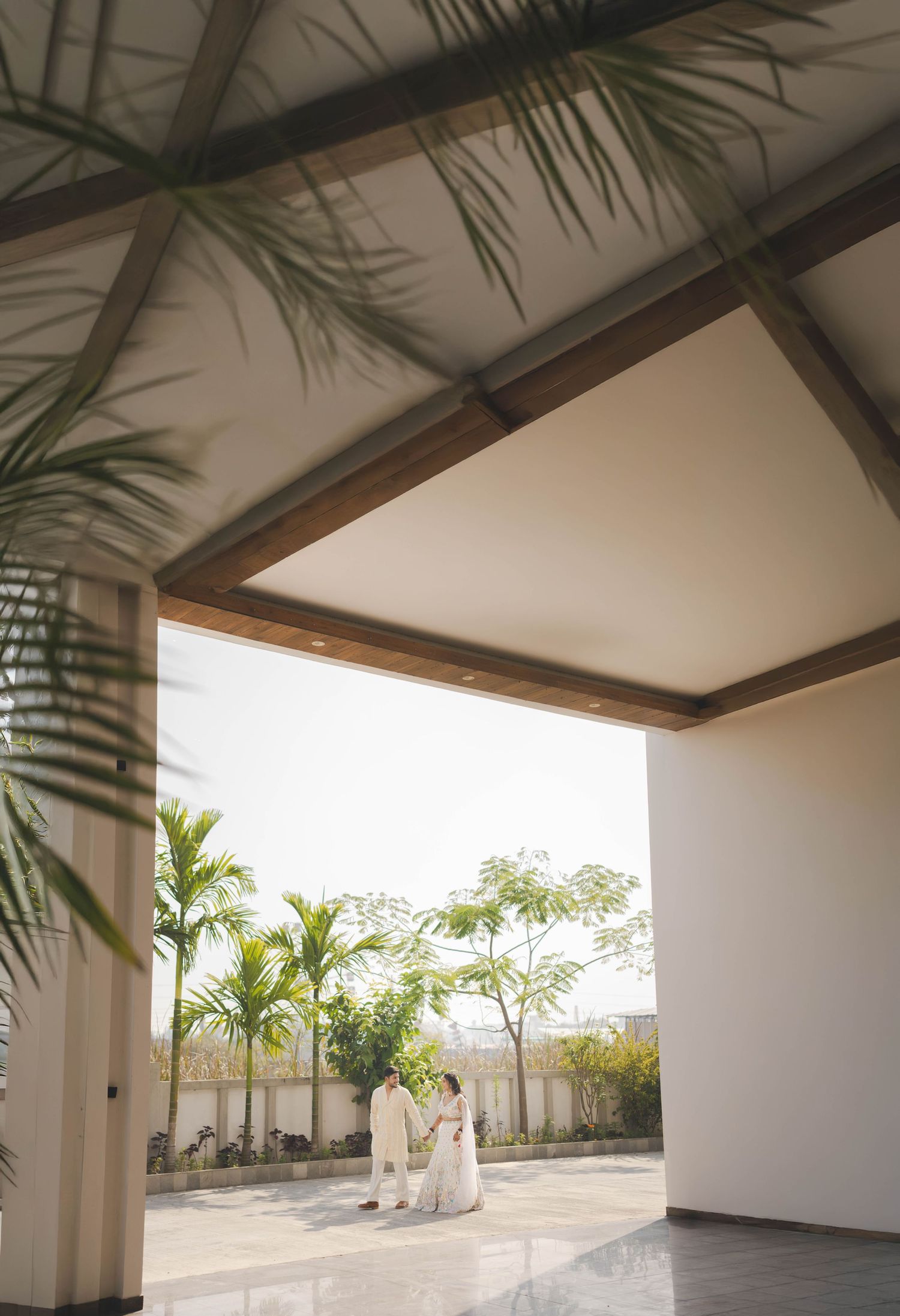 Couple in white attire standing under modern pavilion with wooden beams, surrounded by tropical greenery.