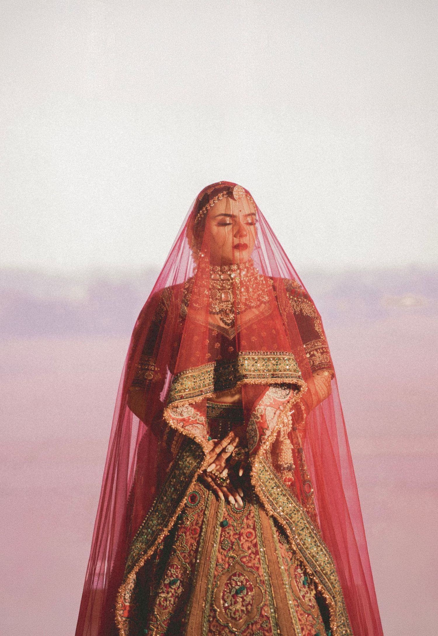 A bride in traditional Indian bridal attire with red lehenga, gold jewelry, and flowing red dupatta veil, eyes closed.