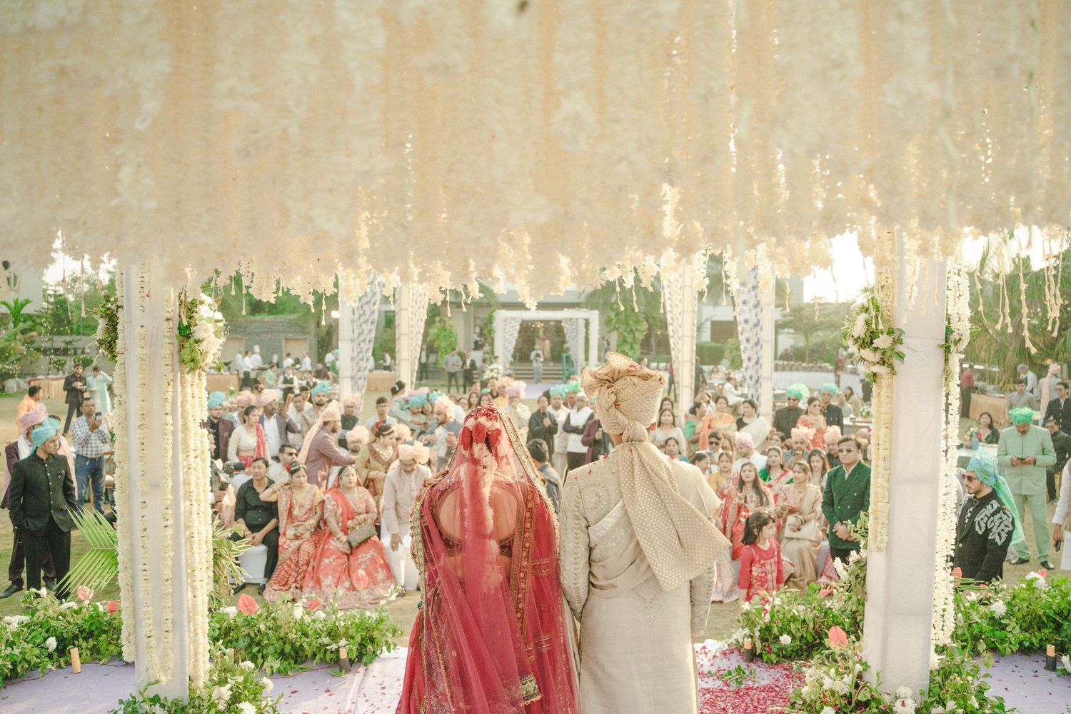 Indian wedding couple stands before floral mandap with guests celebrating at an elegant outdoor ceremony.