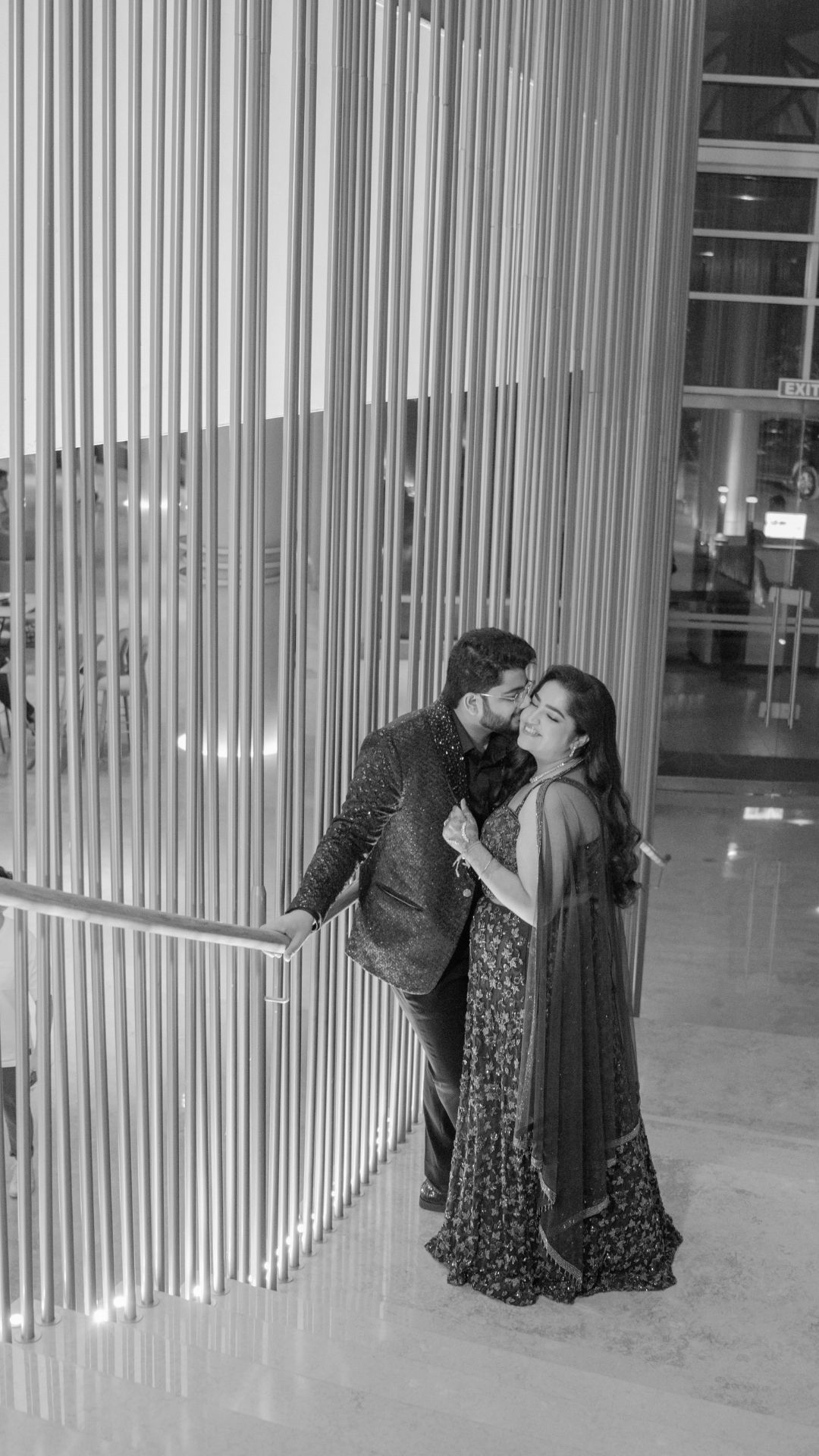 Black and white photo of a couple in traditional Indian attire sharing a romantic kiss in an elegant modern building.