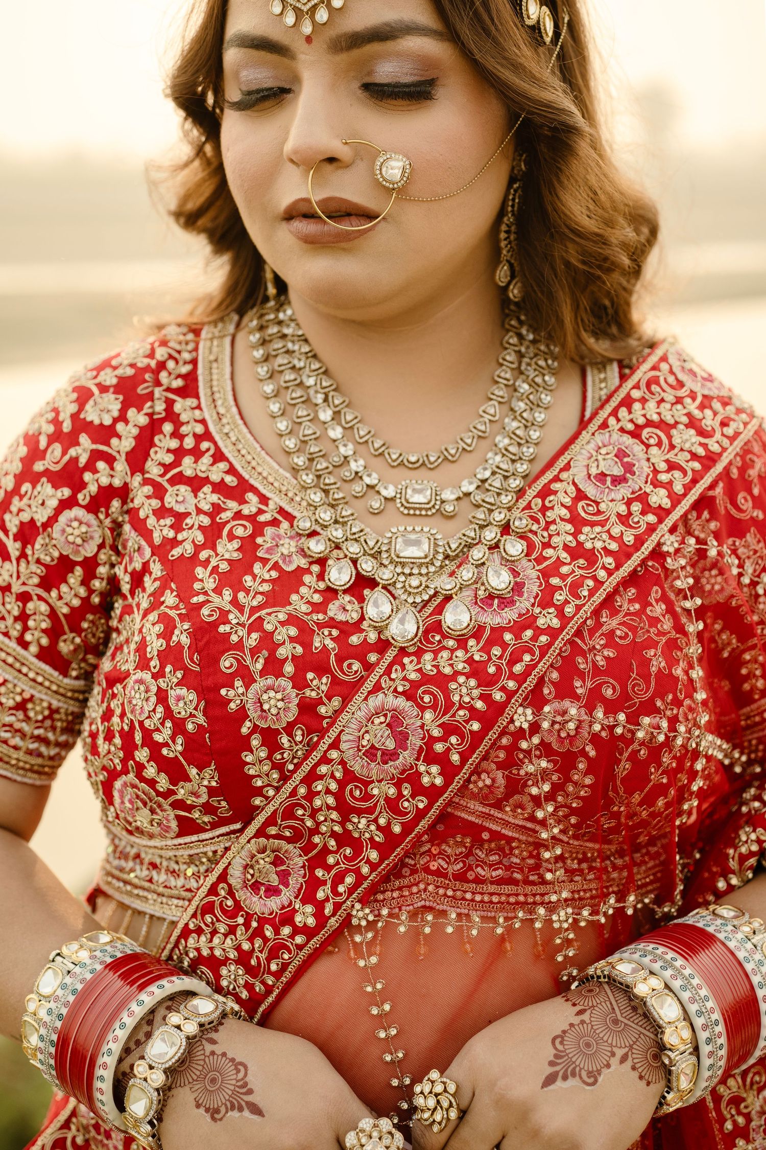 Indian bride in red embroidered lehenga with kundan jewelry, maang tikka, nose ring, and mehndi hands.