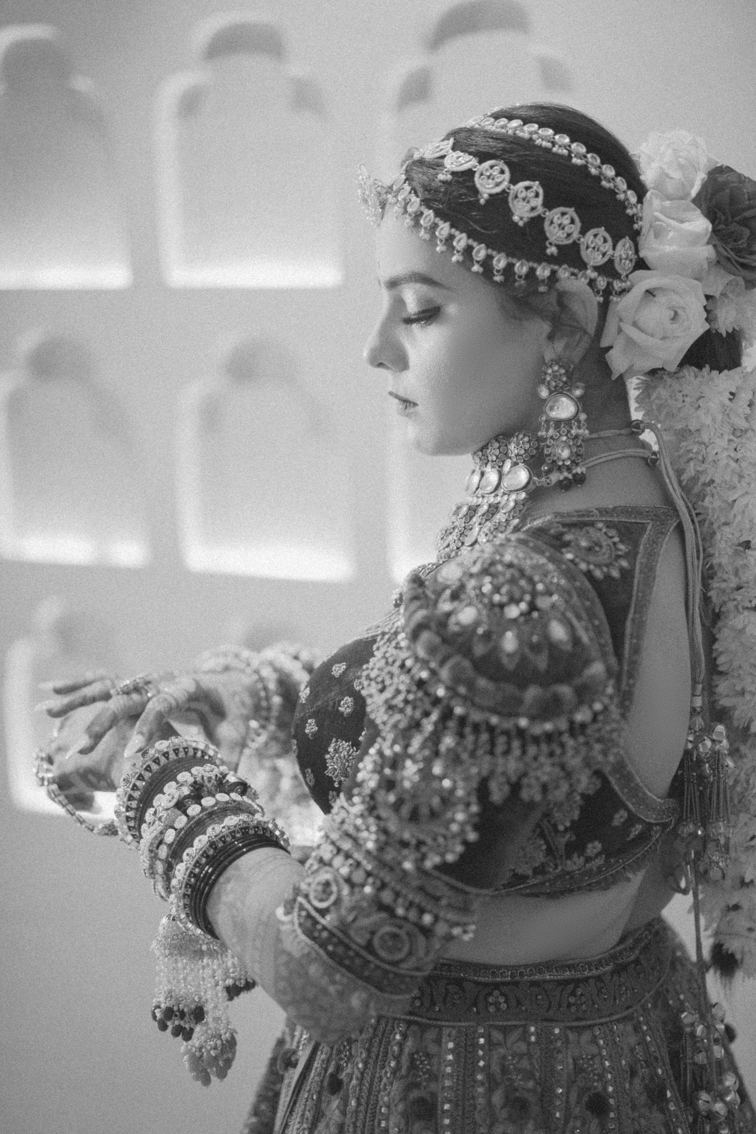 Black and white photo of a bride in traditional Indian bridal jewelry, bangles, and embroidered lehenga with floral hair accessories.