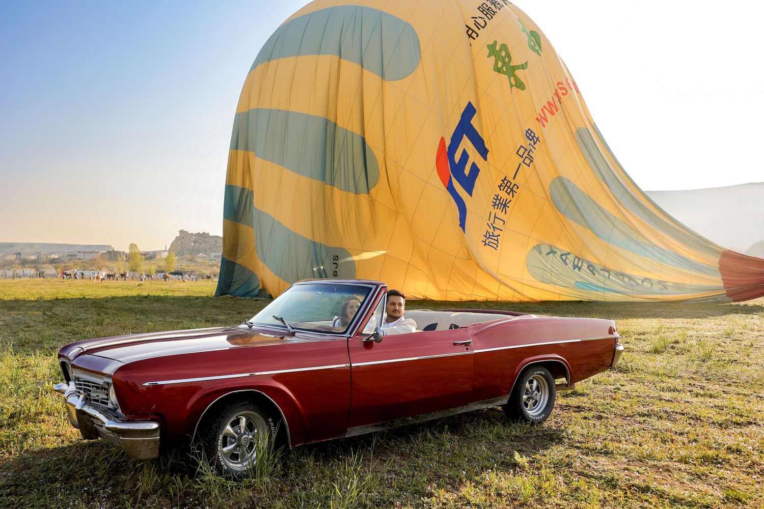 Hot air balloon and vintage red car positioned in grassy field for wedding photos.