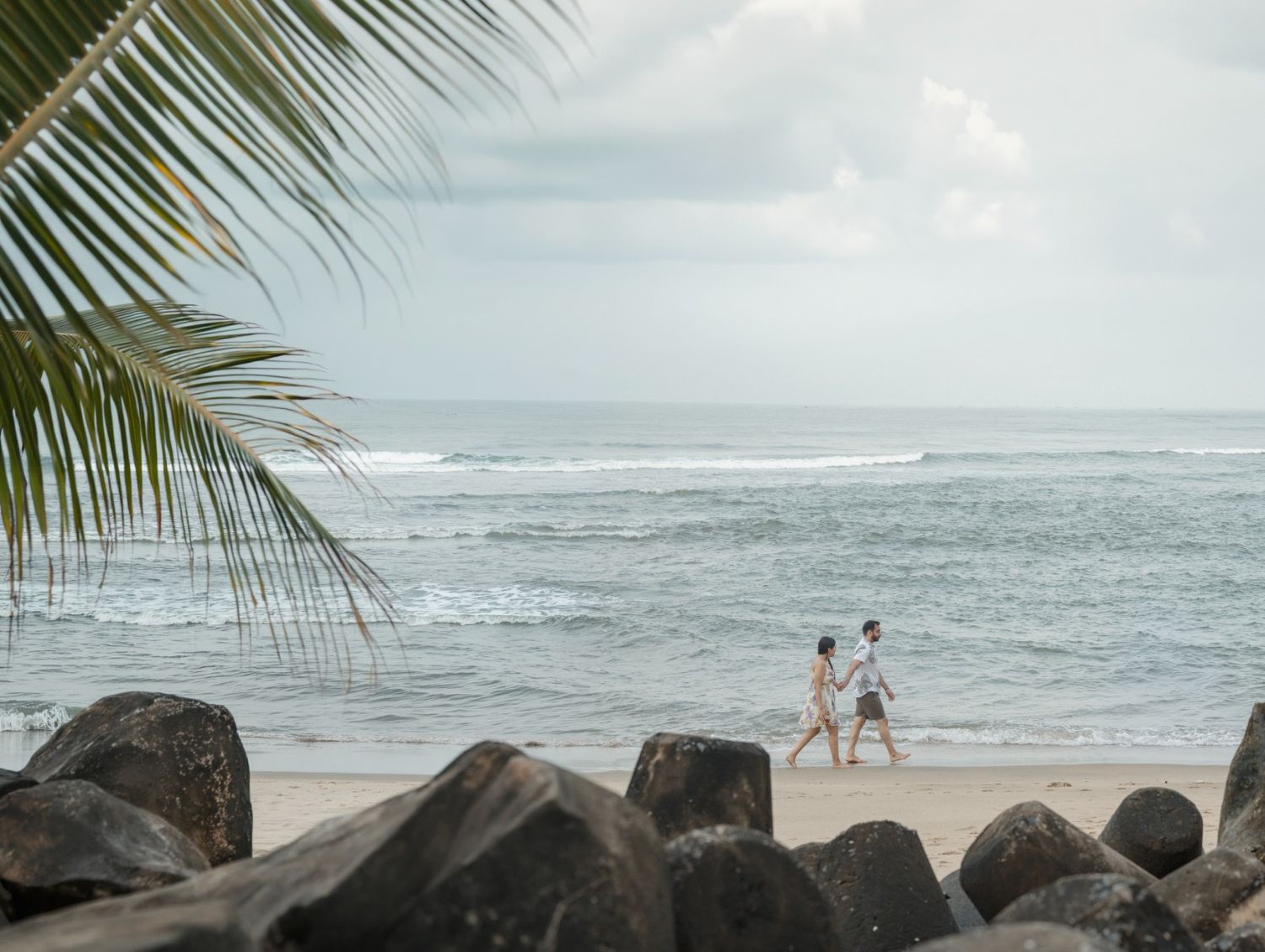 Couple walking hand in hand on a tropical beach with rocky shoreline, palm trees, and ocean waves under cloudy skies.