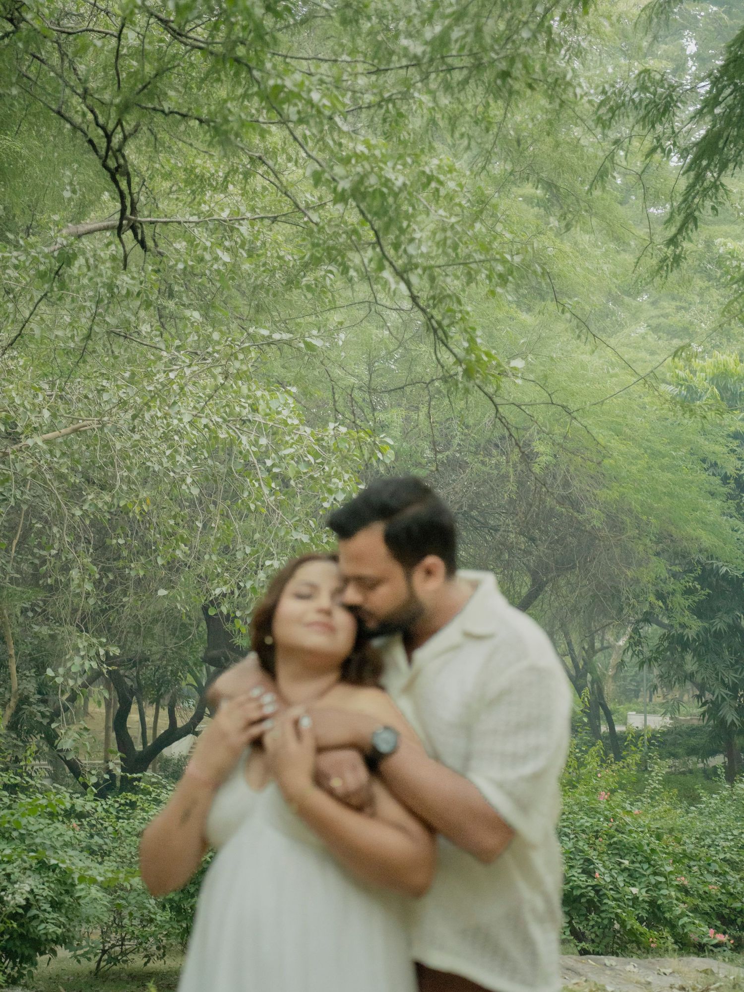 A couple shares a tender embrace surrounded by lush green trees, the man nuzzling the woman from behind in a romantic outdoor setting.