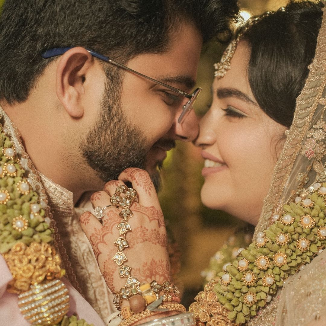 A couple in traditional South Asian wedding attire share an intimate moment, adorned with floral jewelry and mehndi.