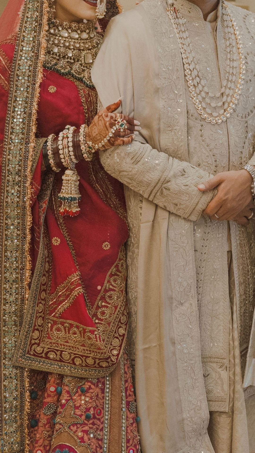 A bride in red embroidered lehenga with gold jewelry holds groom in cream sherwani with pearl mala at South Asian wedding.