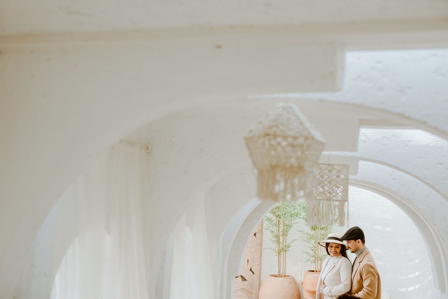 Couple poses romantically under white arched colonnade with macrame decor and soft natural light.